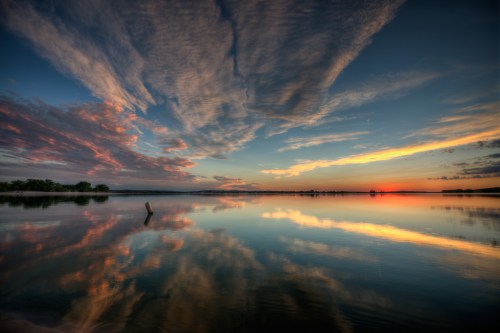 A serene lake at sunset, reflecting colorful clouds and a vibrant sky, with calm water and distant trees.