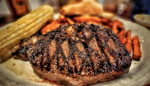 A grilled steak with char marks, served with sweet potato fries and corn on the cob on a white plate.