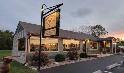 A cozy restaurant exterior at dusk, featuring a sign, large windows, and landscaped surroundings.