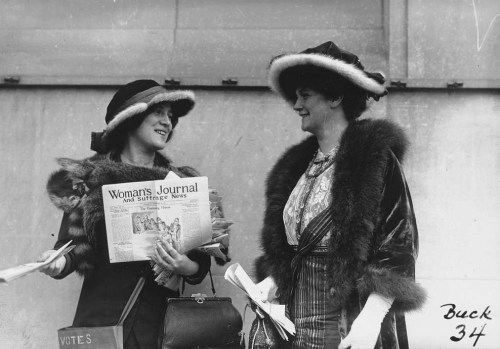 Two women in early 20th-century attire, holding a newspaper titled "Woman's Journal," engaged in conversation.