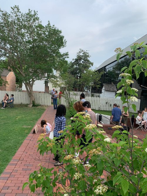 A group of people gathered outdoors in a garden, with greenery and a building in the background.