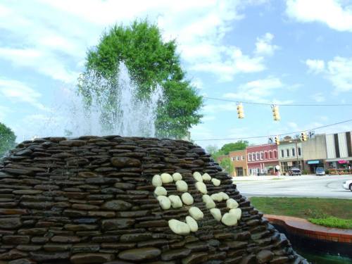 A stone fountain with water spraying, set against a backdrop of a small town street and cloudy sky.