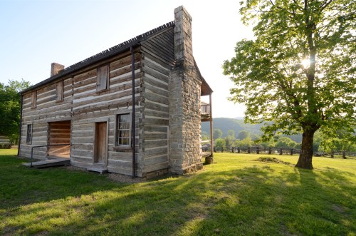 A historic log cabin with a stone chimney, surrounded by green grass and trees, under a bright sky.