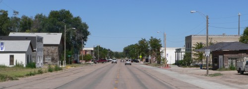 A quiet street in a small town, lined with buildings and trees under a clear blue sky.