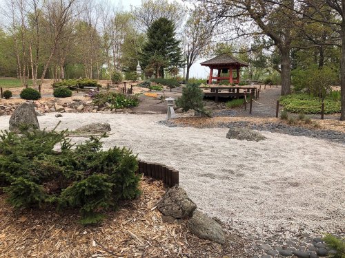 A serene garden featuring gravel paths, rocks, greenery, and a gazebo in the background surrounded by trees.