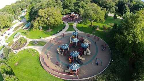 Aerial view of a playground surrounded by green trees and pathways, with children playing on equipment.