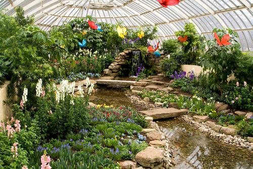 A vibrant greenhouse filled with colorful flowers, a small stream, and butterflies hanging from the ceiling.