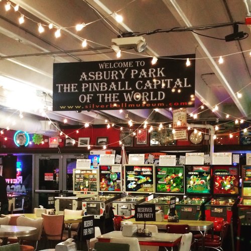A vibrant arcade with pinball machines, string lights, and a sign welcoming visitors to Asbury Park, the Pinball Capital of the World.