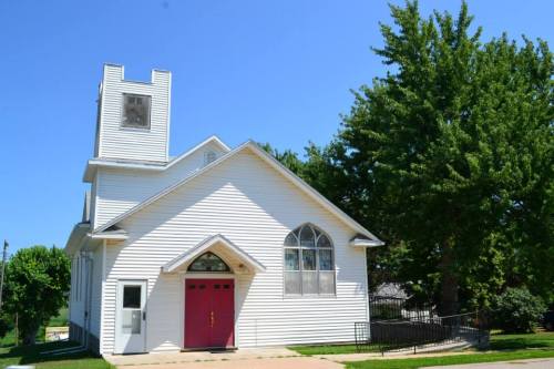 A white church with a red door and a tall tower, surrounded by green trees under a clear blue sky.