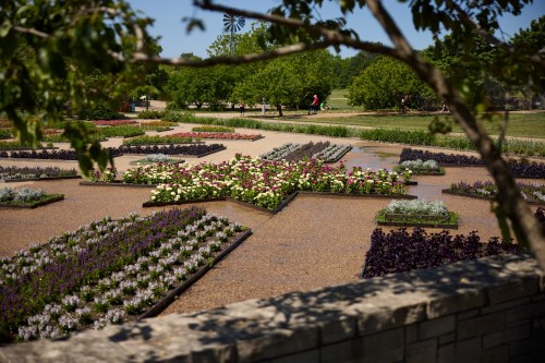 A vibrant garden with colorful flower beds arranged in patterns, surrounded by greenery and a clear blue sky.