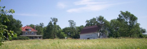 A scenic view of a grassy field with two houses in the background under a clear blue sky.