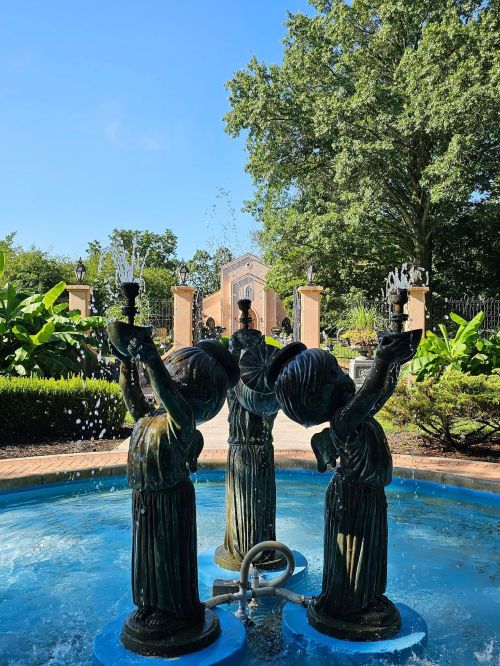 Three bronze statues of children holding torches in a fountain, surrounded by lush greenery and a clear blue sky.