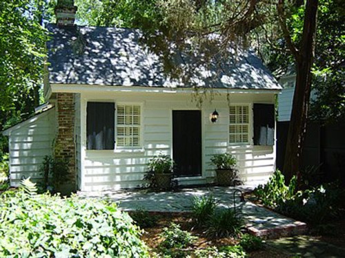 A small white cottage surrounded by greenery, featuring black shutters and a welcoming front porch.