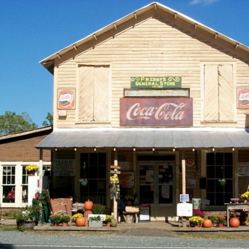 A rustic general store with a Coca-Cola sign, surrounded by pumpkins and flowers, set in a quaint town.