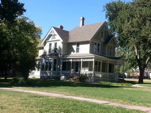 A large, two-story Victorian house with a wraparound porch, surrounded by green grass and trees under a clear blue sky.
