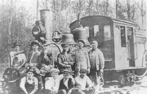 A group of men poses in front of an old steam locomotive in a forested area.