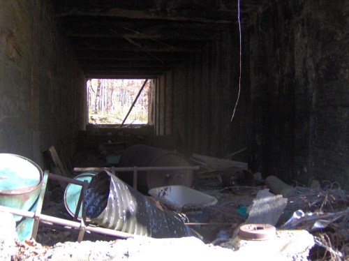 A dark, abandoned tunnel filled with debris, including barrels and a bathtub, leading to a bright opening in the distance.