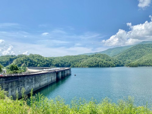 A serene lake surrounded by green hills, with a dam and blue sky dotted with clouds in the background.