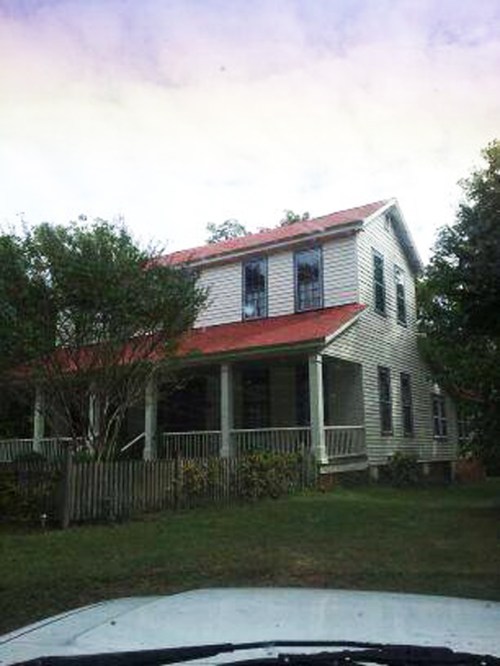 A two-story house with a red roof, white siding, and a porch, surrounded by trees and a grassy yard.