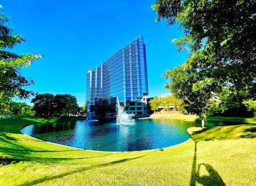 A modern glass building beside a serene pond, surrounded by lush greenery and clear blue skies.