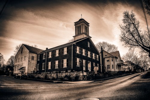Historic brick building with a tower, surrounded by trees, under a dramatic sky, in a sepia-toned landscape.