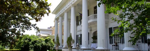 A grand historic mansion with tall columns, surrounded by lush greenery and trees.