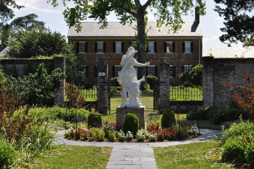 A white statue in a garden, surrounded by flowers and greenery, with a historic building in the background.