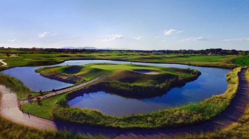 A scenic golf course featuring a green island surrounded by water, with blue skies and lush greenery in the background.