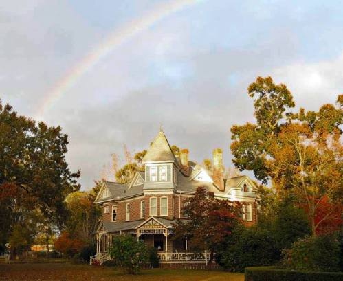A charming Victorian house surrounded by trees, with a rainbow arching across a cloudy sky above.