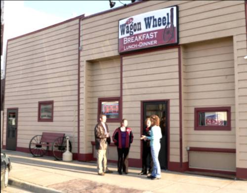 Four people stand outside the Wagon Wheel restaurant, which offers breakfast, lunch, and dinner. The building has a sign above.