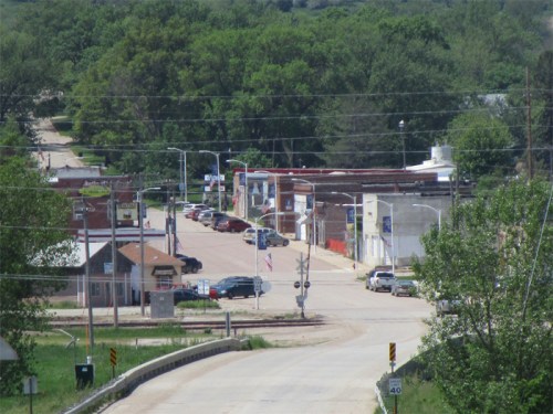 A small town street lined with shops and trees, with cars parked along the road and a clear blue sky above.
