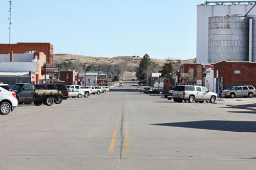 A quiet street lined with buildings and parked cars, leading to a distant hill under a clear blue sky.