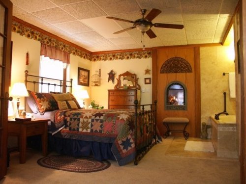 Cozy bedroom with a quilted bed, wooden furniture, and a small fireplace, featuring warm lighting and decorative accents.