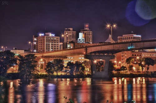 Night view of a city skyline with illuminated buildings reflecting on a river, under a bridge.