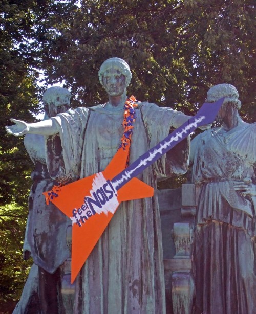 A statue with a large orange guitar decoration, surrounded by two other figures, set in a park.