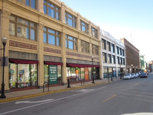 Historic buildings line a street, featuring storefronts with large windows and "For Sale" signs. Clear blue sky above.