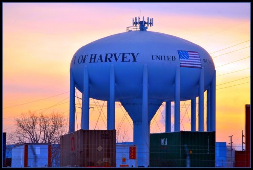 Water tower labeled "City of Harvey" with an American flag, set against a colorful sunset and shipping containers below.