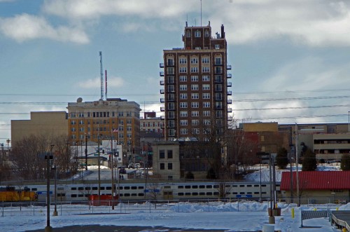 A snowy urban landscape featuring a tall historic building and surrounding structures under a cloudy sky.