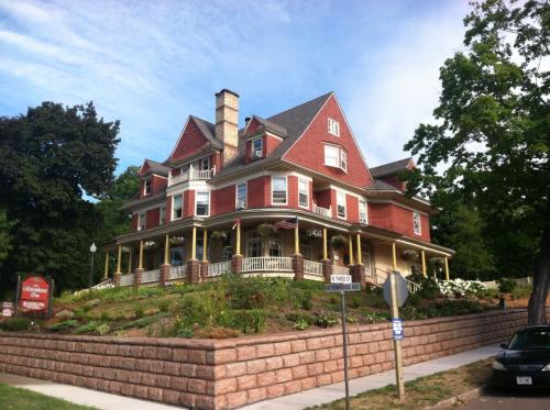 A large, historic red house with a steep roof, surrounded by greenery and flowers, set on a sloped lawn.