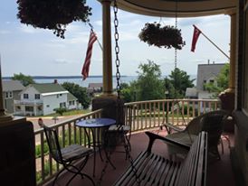A porch with a view of the water, featuring hanging plants, a table, and American flags in the background.