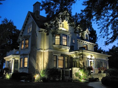 A beautifully lit Victorian-style house at dusk, surrounded by trees and garden, showcasing intricate architectural details.