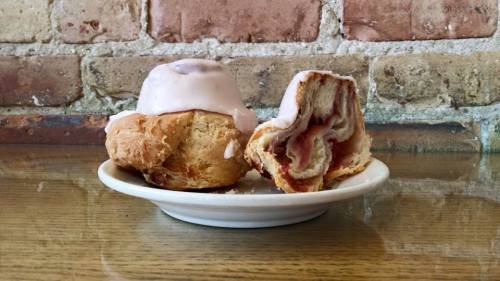 A plate with a glazed pastry, partially sliced to reveal swirls of filling, against a brick wall background.
