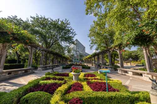 A landscaped garden with colorful flower beds, stone pathways, and a glass greenhouse in the background under a clear sky.