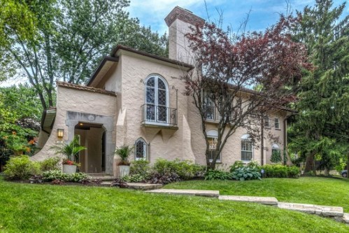 A charming two-story house with a light exterior, large windows, and lush greenery surrounding the front yard.