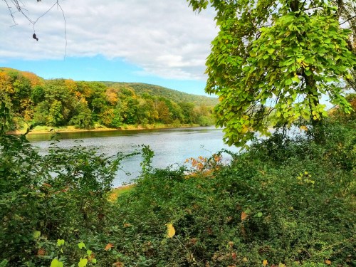 A serene river scene surrounded by lush greenery and colorful autumn trees under a partly cloudy sky.
