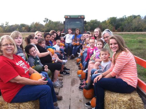 A group of children and adults sitting on a hayride, holding pumpkins, with a tractor in the background.