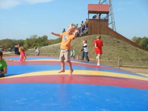 A child jumps joyfully on a colorful inflatable surface, with other kids playing and a wooden tower in the background.