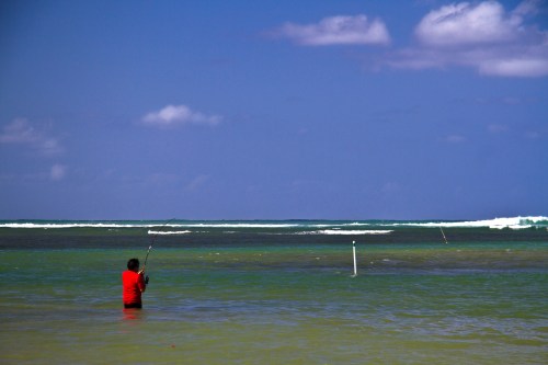 A person in a red shirt stands in shallow water, fishing with a rod against a backdrop of waves and blue sky.