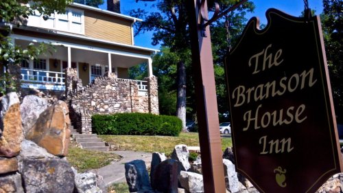 A charming inn with a stone facade and a sign reading "The Branson House Inn," surrounded by greenery.