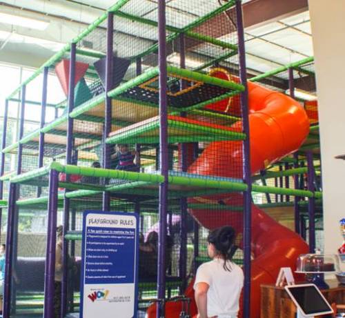 Indoor playground structure with colorful slides and climbing areas, featuring a sign with playground rules in front.
